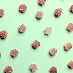 Può includere: Una collezione di piccole perline rosa a forma di rosa sparse su una superficie verde chiaro. Le perline sono scolpite con petali dettagliati, che mostrano un delicato design floreale. Ideale per la creazione di gioielli o progetti artigianali.