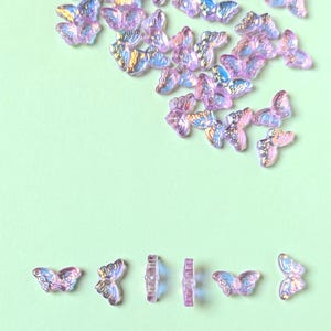 May include: Iridescent butterfly-shaped embellishments in shades of purple, pink, and blue are scattered across a light green background. The decorative elements are arranged in a random pattern, with some lying flat and others standing upright.