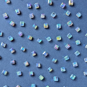 Può includere: Una dispersione di perline iridescenti, tagliate a forma quadrata, su una superficie blu scuro. Le perline riflettono uno spettro di colori, tra cui blu, verde e viola. Piccole, adatte per la creazione di gioielli o artigianato.