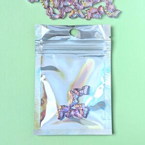 May include: A clear, iridescent bag containing several small, purple butterfly-shaped embellishments. Additional butterflies are scattered above the bag on a light green background. These decorative elements could be used for crafts or nail art.