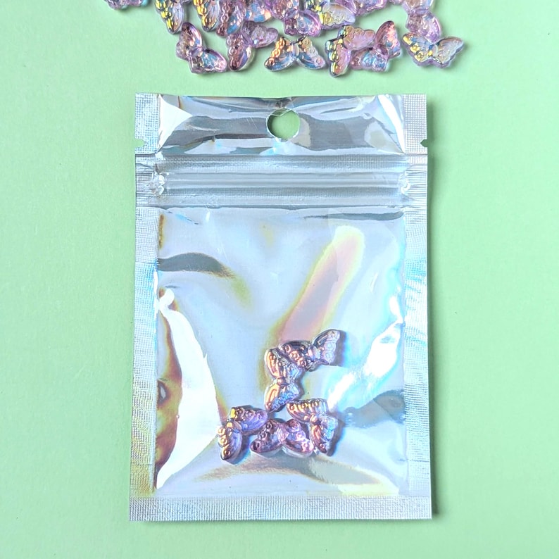 May include: A clear, iridescent bag containing several small, purple butterfly-shaped embellishments. Additional butterflies are scattered above the bag on a light green background. These decorative elements could be used for crafts or nail art.