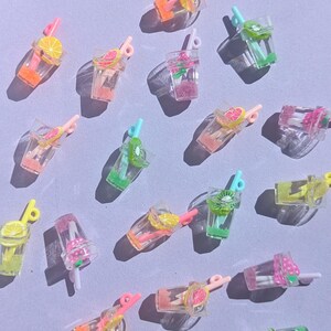 Peut inclure: Une collection de breloques miniatures en plastique transparent représentant des boissons avec des tranches de fruits colorées et des pailles. Les breloques présentent des fruits orange, roses, verts et jaunes, dont des citrons, des fraises et des kiwis.