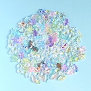 Può includere: Una collezione di abbellimenti artigianali iridescenti e traslucidi in varie forme, tra cui stelle marine, conchiglie e pesci. I colori vanno dal rosa e viola pastello al blu e al verde. Gli oggetti sono sparsi su uno sfondo azzurro chiaro.