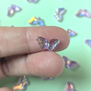 May include: A small, iridescent butterfly-shaped embellishment with a clear body and detailed wing patterns. The butterfly is held between fingers, with other similar embellishments scattered on a light green surface.