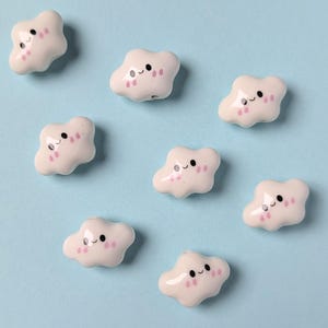 Può includere: Perline bianche a forma di nuvola, con faccine sorridenti e dettagli rosa, disposte su uno sfondo azzurro chiaro. Ogni perlina ha un foro per l'infilatura, perfette per lavori creativi.
