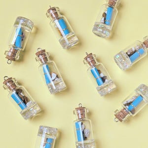 Könnte beinhalten: Eine Sammlung kleiner Glasflaschen mit Korken. Jede Flasche enthält eine aufgerollte blaue Papierrolle und kleine dekorative Elemente wie Kieselsteine oder Muscheln. Die Flaschen haben einen transparenten, strukturierten Boden und eine kleine Metallschlaufe.