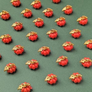 Op de afbeelding: Een verzameling kleine, oranje pompoenvormige bedels gerangschikt in een rasterpatroon. Elke bedel heeft een feloranje, geribbelde pompoen met een gestructureerd, goudkleurig bladaccent en een klein lusje om hem te bevestigen.