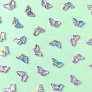 May include: A collection of iridescent butterfly-shaped embellishments scattered on a light green background. The butterflies have a translucent appearance with blue and yellow accents on their wings, creating a shimmering effect. These decorative elements are ideal for crafts.