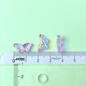 May include: Three iridescent butterfly-shaped craft embellishments. The butterflies are pink and blue, with a clear, textured appearance. A ruler with centimeter markings is in the foreground. The background is a light green color.