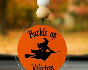 Op de afbeelding: Oranje auto luchtverfrisser met een geschulpte rand, met een zwart silhouet van een heks op een bezemsteel en de woorden "Buckle up Witches". De luchtverfrisser wordt opgehangen door een zwart koord en houten kralen.