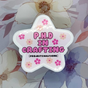 Könnte beinhalten: Ein sternförmiger Aufkleber mit dem Text "P.H.D IN CRAFTING" in rosa Buchstaben, umgeben von rosa und pfirsichfarbenen Blumengrafiken. Der Aufkleber enthält auch den Text "(PROJECTSHALFDONE)" in einer kleineren Schriftart. Der Hintergrund zeigt ein Blumenmuster.
