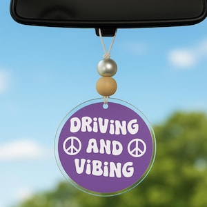 May include: A car charm hanging from a rearview mirror. The charm has a purple circular design with the words "DRIVING AND VIBING" and peace symbols in white. Above the charm are three beads in silver and wood tones.
