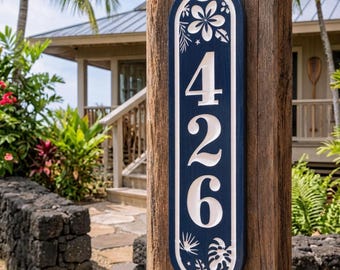 May include: A vertical, oval-shaped house number sign featuring the digits 426 in white against a dark blue background. The sign is decorated with carved white floral and tropical leaf motifs at the top and bottom, and is mounted on a wooden post.