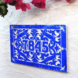 May include: A vibrant blue rectangular house number plaque with the number "7845" in white. The plaque features an intricate, openwork design with floral and leaf patterns. The background is a textured white wall, with pink flowers in the upper portion of the image.