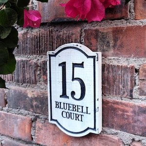May include: A white address plaque with the number 15 and the text "BLUEBELL COURT" in black lettering, mounted on a red brick wall. Pink flowers and green leaves are visible in the background.