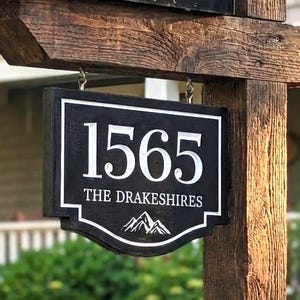 May include: A black metal mailbox with a red flag mounted on a rustic wooden post. A rectangular black sign hangs from the post, featuring the white text 1565, THE DRAKESHIRES, and a small mountain graphic at the bottom of the sign.
