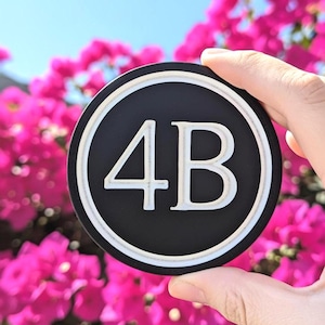 May include: A black and white round coaster with the numbers "4B" in white. The coaster has a white border and is held in front of a background of pink flowers and a blue sky.