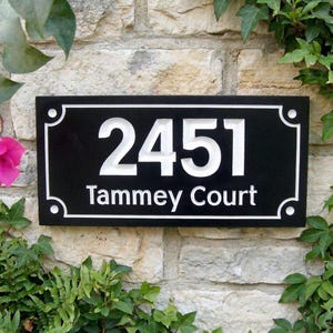 May include: A black rectangular house number sign with white detailing. The number "2451" is displayed above the words "Tammey Court". The sign is surrounded by foliage and mounted on a stone wall.