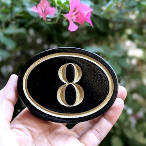 May include: Oval black house number eight plaque with a gold border and gold number. The plaque is held in a hand against a blurred background of green foliage and pink flowers.