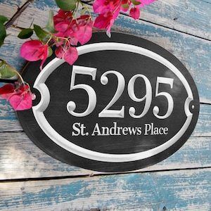 May include: Oval-shaped address plaque with the number "5295" and "St. Andrews Place" in white lettering on a black background. The plaque has a white border and is set against a weathered blue wooden surface. Pink flowers are in the upper left corner.