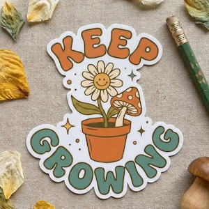 May include: A sticker with the words "KEEP GROWING" in orange and teal lettering. The sticker features a smiling daisy and a mushroom in a terracotta pot. Dried flower petals and a pencil are scattered around the sticker.