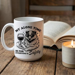 以下が含まれることがあります： サングラスをかけたアナグマがワイングラスを持っている黒いグラフィックが描かれた白いセラミックマグ。マグには「My Alone Time Is For Everyone's Safety」と書かれています。マグは木製の表面に置かれています。