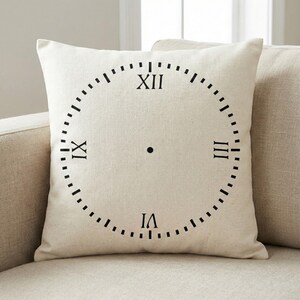 Puede incluir: Un cojín cuadrado de color crema con un diseño de esfera de reloj negro. La esfera del reloj tiene números romanos y marcadores de minutos. El cojín está sobre un sofá beige.