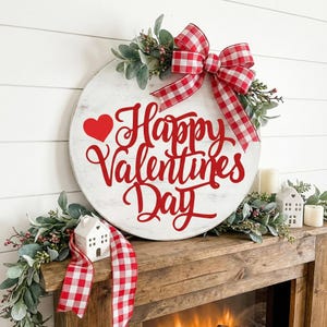 May include: A round, white wooden sign with the text "Happy Valentine's Day" in red script, accented with a red heart. The sign is decorated with a red and white checkered bow, greenery, and a matching ribbon. The sign is displayed on a wooden mantel.