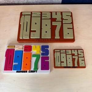 May include: Three number puzzles with the text "NUMBER CRAFT". The puzzles are in various colors, including red, blue, orange, green, and purple. The numbers 1, 0, 9, 8, 7, and 6 are featured. The puzzles are on a light wooden surface.