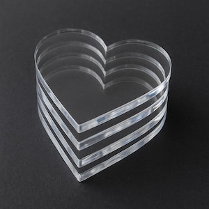 May include: A stack of clear, heart-shaped acrylic pieces. The hearts are layered, creating a three-dimensional effect. The edges are smooth and the material is transparent, allowing light to pass through. The background is a solid black.