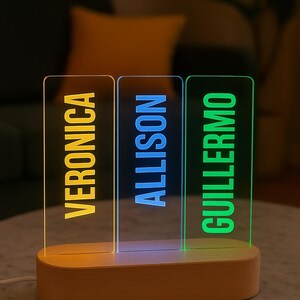 May include: A personalized LED night light with three acrylic panels displaying the names "Veronica," "Allison," and "Guillermo" in yellow, blue, and green, respectively. The names are illuminated against a clear background, set on a wooden base.