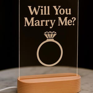 Può includere: Un'insegna in acrilico trasparente con la scritta "Will You Marry Me?" e un disegno di anello. L'insegna illuminata è posta su una base di legno chiaro. Un cavo bianco esce dalla base. Ideale per una proposta di matrimonio.