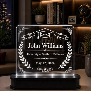 May include: A personalized graduation gift featuring a glowing acrylic plaque with the name "John Williams", a graduation cap, and scrolls. The plaque includes "University of Southern California" and "May 12, 2024". The base is black.