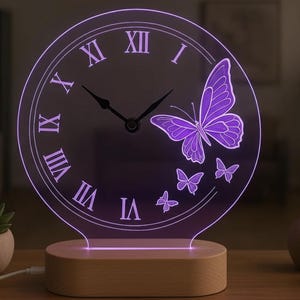 May include: A purple LED clock with Roman numerals and a butterfly design. The clock is made of clear acrylic and sits on a wooden base. The clock's hands are black, and the butterflies are a lighter shade of purple.
