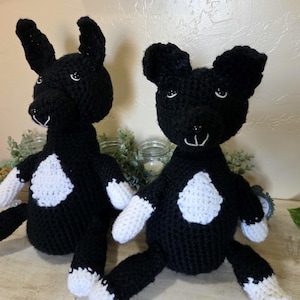 May include: Two handmade black and white crochet dog stuffed animals. The dogs have black bodies, white patches on their chests, and white paws. One dog has upright ears, and the other has floppy ears. They are sitting on a light-colored surface.
