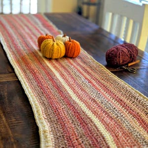 May include: A hand-crocheted table runner with a striped pattern in shades of red, brown, and cream. The runner is decorated with four small crocheted pumpkins in orange, yellow, and white. A ball of yarn and scissors are also on the table.