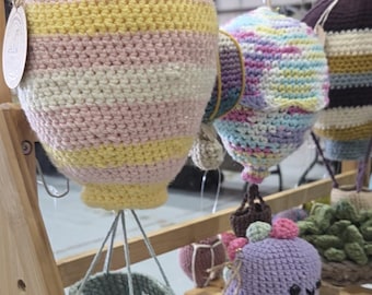 May include: Crocheted hot air balloon-shaped hanging planters in pastel colours, with a matching hanging basket below. Other crocheted items, including a purple creature, are displayed on a wooden shelf. The balloons are suspended from the ceiling.