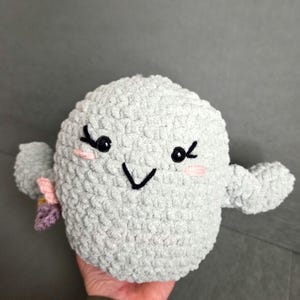 Puede incluir: Un juguete con forma de fantasma de crochet de felpa gris claro, con ojos negros, pestañas y una boca sonriente. El fantasma tiene brazos pequeños y rubor rosa en las mejillas. Una pequeña flor rosa y morada está unida al costado.