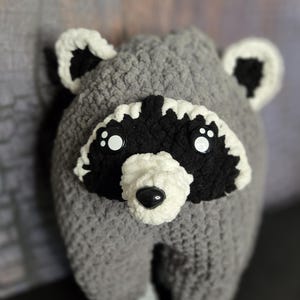 May include: A plush raccoon toy crafted from soft grey yarn, featuring a prominent black and white facial mask, a white muzzle with a black plastic nose, and round black eyes with white accents. The ears are white with black centres.