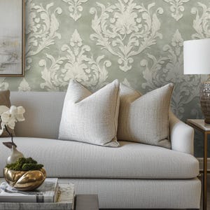 May include: A light gray sofa with two light beige throw pillows. The background features a sage green and white damask wallpaper. A gold-toned decorative bowl and a white lamp are also visible.