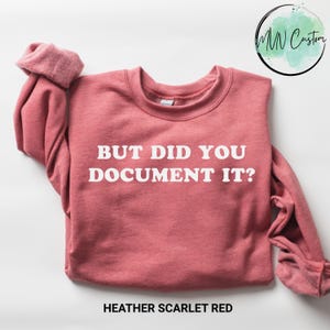 May include: A heather scarlet red crewneck sweatshirt featuring the white text "BUT DID YOU DOCUMENT IT?" printed across the chest. The garment has long sleeves with ribbed cuffs and a ribbed neckline, showcasing a soft, fleece-lined interior texture.