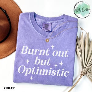 May include: A violet short-sleeve t-shirt with a crew neckline featuring the white text Burnt out but Optimistic and four-pointed star graphics. The shirt has a Comfort Colors label at the inner neck and is made of a soft, pigment-dyed cotton fabric.