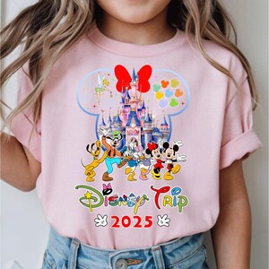 May include: Pale pink t-shirt with a vibrant Disney-themed graphic. The design includes Mickey and Minnie Mouse, a castle, and the text "Disney Trip 2025". The overall design is bright and cheerful.