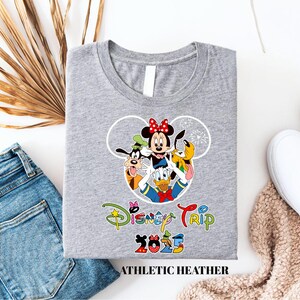 May include: A grey t-shirt with a Disney-themed graphic. The design includes Minnie Mouse, Goofy, Donald Duck, and Pluto. The words "Disney Trip 2025" are printed below the characters. The shirt is labelled "ATHLETIC HEATHER".