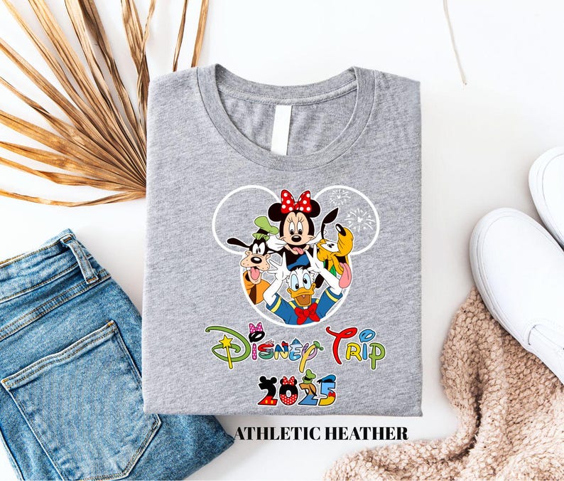 May include: A grey t-shirt with a Disney-themed graphic. The design includes Minnie Mouse, Goofy, Donald Duck, and Pluto. The words "Disney Trip 2025" are printed below the characters. The shirt is labelled "ATHLETIC HEATHER".