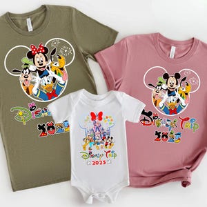 May include: Three Disney-themed tops and a babygrow. The tops are olive green and dusty rose, featuring Mickey Mouse, Minnie Mouse, Donald Duck, Goofy, and Pluto. The babygrow is white with a castle graphic. All items have "Disney Trip 2025" text.