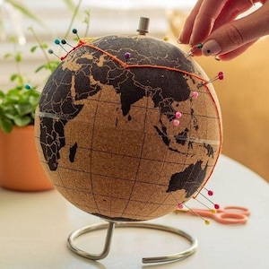 May include: A cork globe with a world map design, mounted on a silver-colored stand. Colorful pins mark locations, connected by orange thread. A hand is placing a pin. Scissors and thread are on the table.