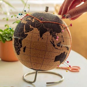 May include: A cork globe with a dark map design, mounted on a silver metal stand. Colorful pins mark locations, connected by orange thread. A hand is placing a pin. Scissors and thread are on the table.