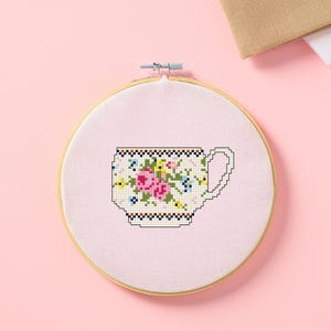 May include: Cross-stitch embroidery of a teacup with pink roses, blue and yellow flowers, and a black and white border. The embroidery is set in a wooden hoop against a pink background. Additional fabric is visible in the background.