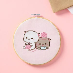 May include: A cross-stitch embroidery hoop featuring two cartoon bear characters. One white bear is holding a pink item, and the other brown bear has a pink ball on its head. The background is light pink.